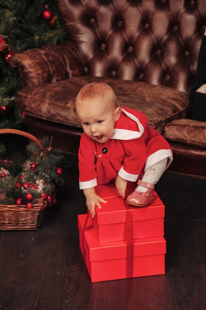 Criança de olhos azuis de um ano de idade com fantasia de Natal, brincando na sala de estar com caixas de árvore e presentes. Bebê fofo com emoção à noite de férias. Conceito de celebração familiar de Natal e Feliz Ano Novo