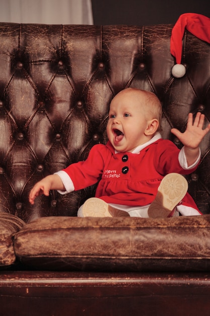 Criança de olhos azuis de um ano de idade em traje de Natal na poltrona na sala de estar. Emoção de bebê na noite de feriado da cadeira antiga. Conceito de celebração familiar de Natal e Feliz Ano Novo