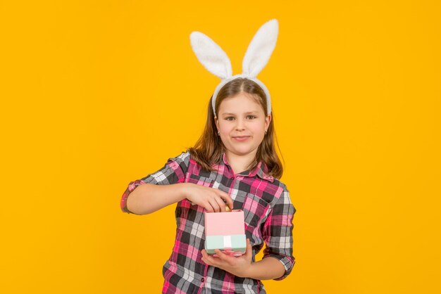 Criança de páscoa em orelhas de coelho abre caixa de presente em fundo amarelo