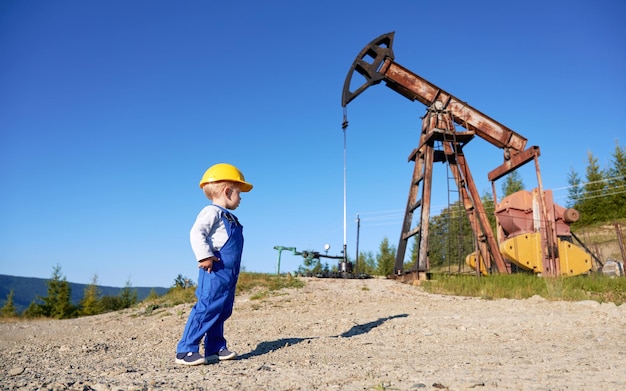 Criança de pé em roupas de trabalho e capacete amarelo protetor na superfície de pedra Menino assistindo a bomba de óleo trabalhando no fundo do céu azul limpo