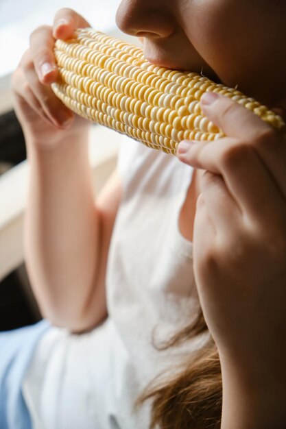 Foto criança de perto a morder um grande vegetal milho em casa comida vegetariana fresca vitaminas nutrição saudável sem ogm dentro de casa rosto irreconhecível