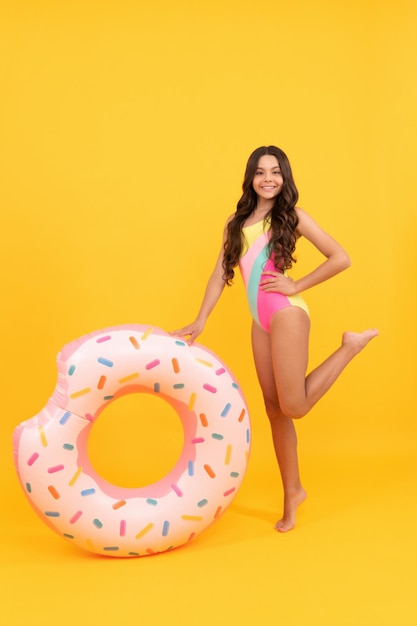 Criança de praia feliz com cabelo encaracolado em traje de banho com anel inflável de rosquinha para diversão de festa na piscina nas férias de verão no parque aquático de fundo amarelo