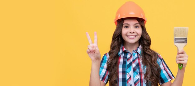 Criança de renovação feliz adolescente em capacete de construção segura pincel de pintura mostra gesto de paz