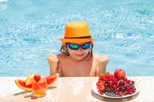 Criança de verão à beira da piscina comendo frutas e bebendo coquetel de limonada Conceito de férias para crianças de verão Garotinho relaxando em uma piscina se divertindo durante as férias de verão