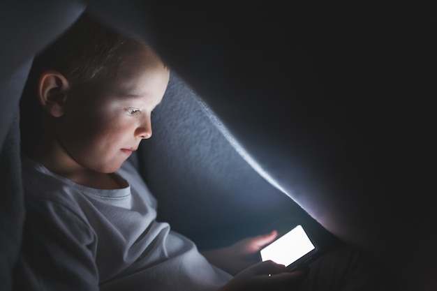 criança debaixo de um cobertor brincando com um smartphone, sentado na Internet. conceito de passar o tempo em isolamento seguro.