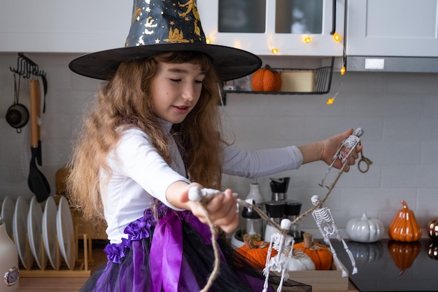Criança decora a cozinha em casa para o Halloween Menina em uma fantasia de bruxa brinca com a decoração para o feriado morcegos jack lanterna abóboras Conforto de outono em casa loft cozinha escandinavo