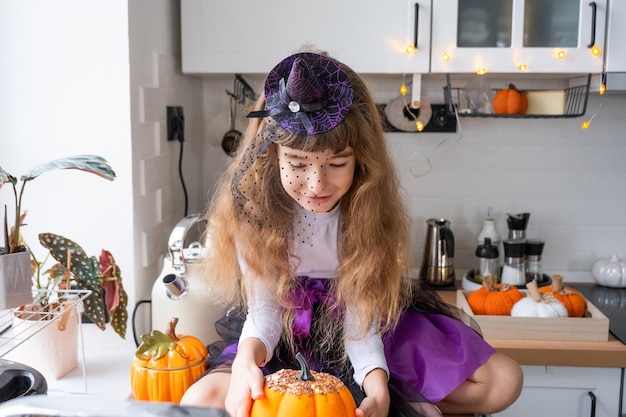 Criança decora a cozinha em casa para o Halloween Menina em uma fantasia de bruxa brinca com a decoração para o feriado morcegos jack lanterna abóboras Conforto de outono em casa loft cozinha escandinavo