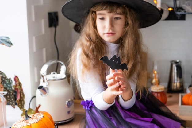 Criança decora a cozinha em casa para o Halloween Menina em uma fantasia de bruxa brinca com a decoração para o feriado morcegos jack lanterna abóboras Conforto de outono em casa loft cozinha escandinavo