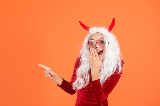 Criança diabo do Dia das Bruxas com chifres de diabinho usando óculos de festa e apontando o dedo no espaço da cópia feliz