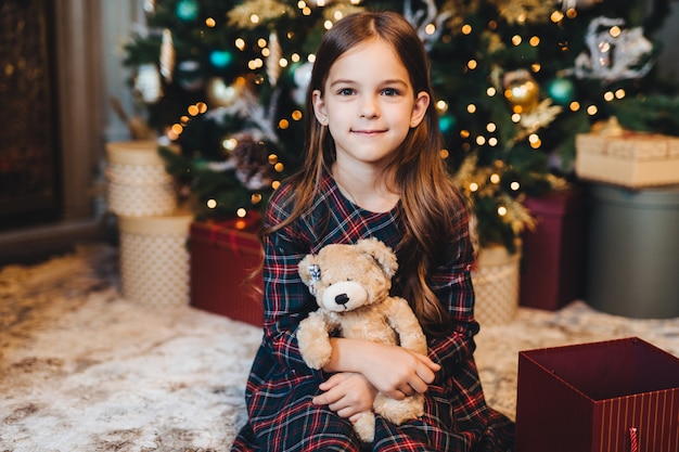 Criança do sexo feminino pequena com aparência atraente, segura o ursinho de pelúcia, recebe presentes dos pais, senta-se perto da árvore de Natal