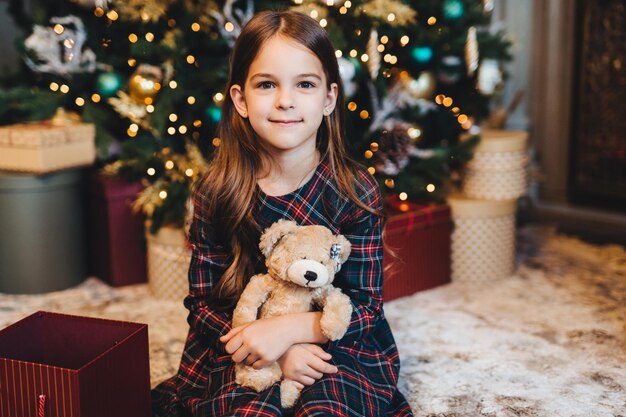 Criança do sexo feminino pequena com aparência atraente segura ursinho de pelúcia recebe presente dos pais se senta perto da árvore de Natal Garota adorável que fica feliz em receber presente no Natal de Ano Novo