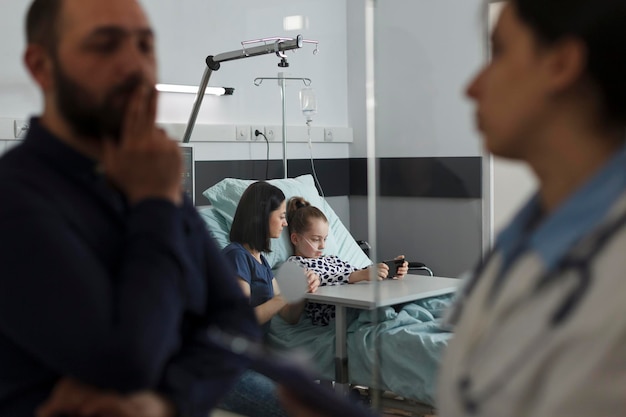 Criança doente hospitalizada assistindo desenhos no celular enquanto descansava na enfermaria pediátrica do hospital. Menina doente jogando no smartphone com a mãe enquanto o pai fala com o pediatra especialista.