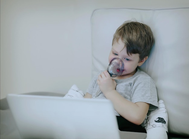 Criança doente sorridente com nebulizador pediátrico olhando para a tela do laptop Criança com asma ou bronquite tem dificuldade para respirar. Menino doente se divertindo navegando na internet ou assistindo a um filme.
