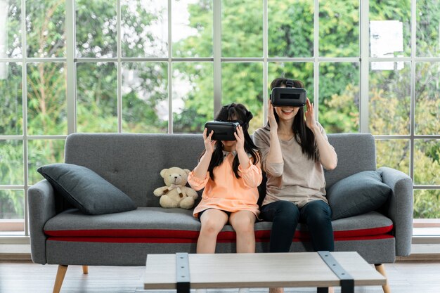 Criança e mulher com fone de ouvido de realidade virtual sorrindo enquanto estão sentadas no sofá dentro de casa