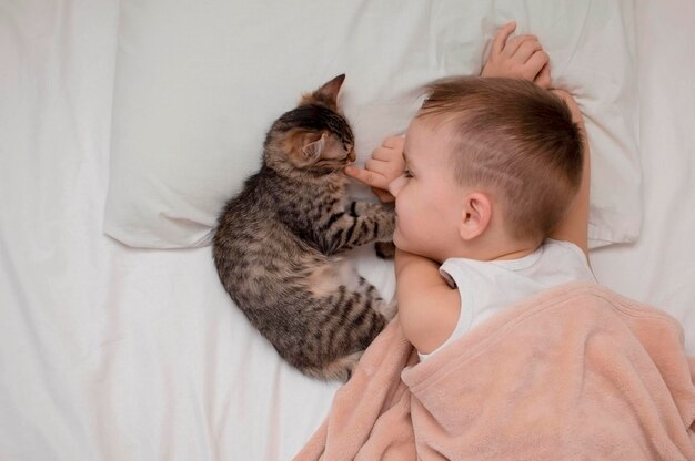 Criança e um gato cinza dormindo juntos em um lençol branco a amizade de infância de um gatinho e uma criança O conceito de animais de estimação