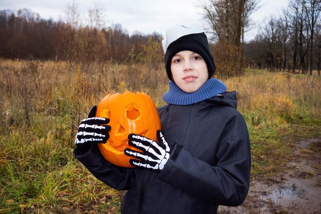 Foto criança em luvas de esqueleto segurando jacko'lantern de abóbora de halloween no fundo das árvores