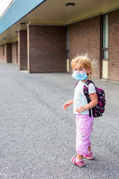 Foto criança em máscara com mochila durante o vírus corona perto de escola ou jardim de infância