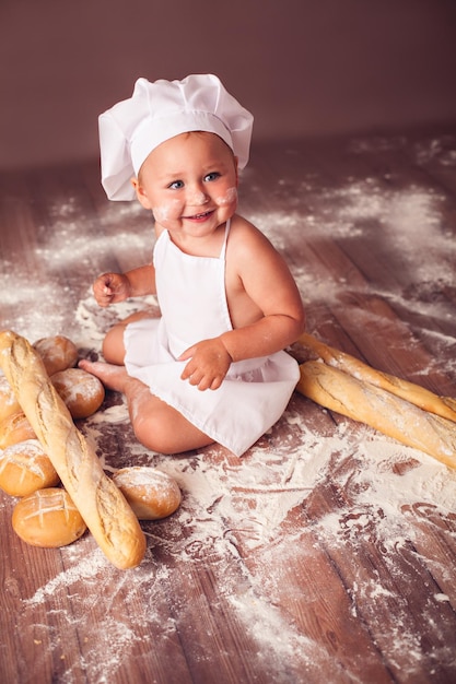 Criança encantadora com chapéu de cozinheiro e avental sentado na farinha com pães rindo alegremente