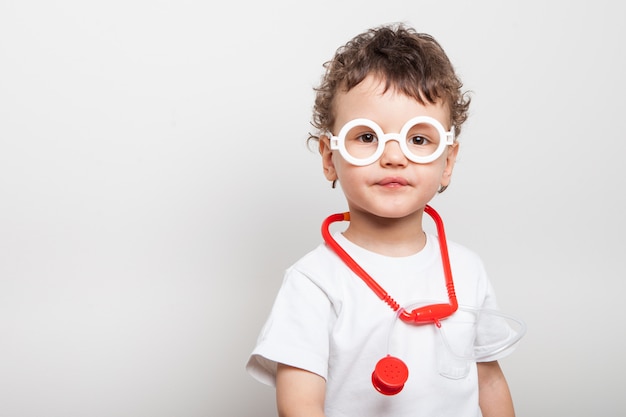 Criança encaracolada bonita e engraçada em um terno de médicos com um estetoscópio no pescoço e nos copos de seu conjunto de brinquedos médico para crianças.