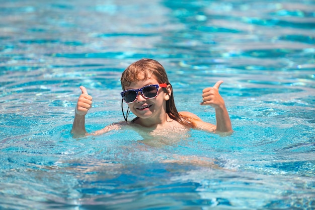 Criança engraçada animada com o polegar para cima em óculos de sol na piscina no dia de verão Criança na piscina de verão