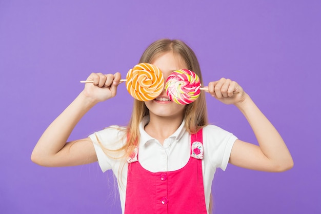 Criança engraçada com pirulitos em fundo violeta. Sorriso de criança com doces em varas no fundo roxo. Menina sorrindo com caramelos de redemoinho. Comida e sobremesa. Beleza com doces. Dieta e dieta.