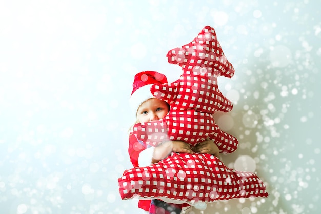 criança engraçada em um terno de Papai Noel abraça uma árvore de Natal têxtil vermelha