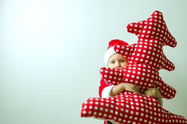 criança engraçada em um terno de Papai Noel abraça uma árvore de Natal têxtil vermelha