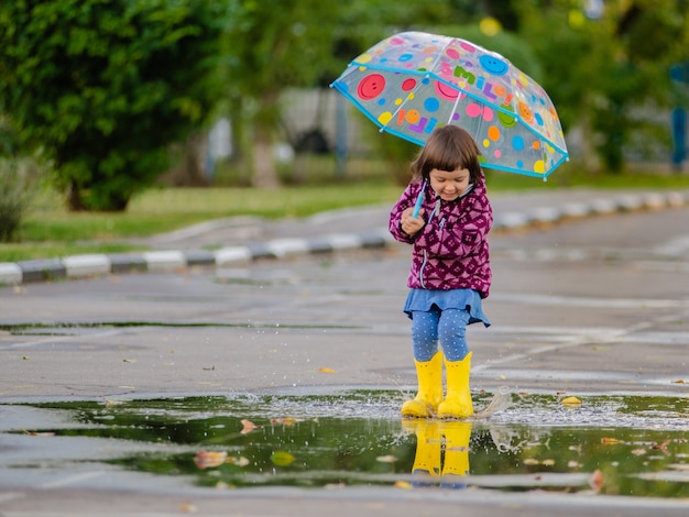 Criança engraçada feliz com guarda-chuva multicolorida pulando poças em botas de borracha e rindo