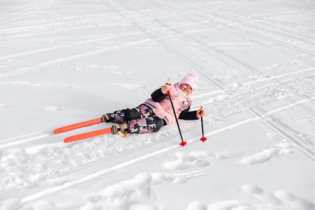 Foto criança está deitada na neve menina feliz em um terno rosa está aprendendo a esquiar e caiu na neve da estrada de neve ...