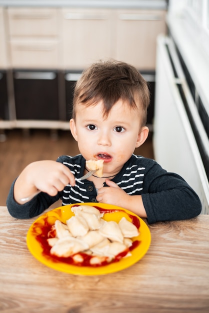 Foto criança faminta comendo bolinhos na cozinha, sentada à mesa com uma jaqueta cinza