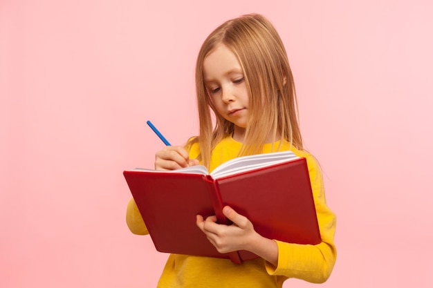 Criança fazendo lição de casa. retrato de menina inteligente escrevendo poema em livro com expressão concentrada séria, desenvolvimento de habilidades criativas de criança. tiro de estúdio interior isolado no fundo rosa