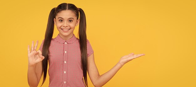 Criança feliz apresentando produto em fundo amarelo espaço de cópia mostrar ok anunciar rosto de criança cartaz horizontal menina adolescente retrato isolado bandeira com espaço de cópias