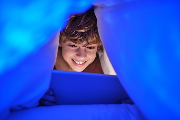 Criança feliz assistindo vídeo no tablet com um sorriso enquanto está deitada debaixo de um cobertor no quarto com luz fraca em casa à noite