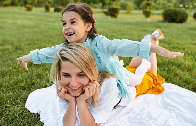 Criança feliz brincando com a mãe bonita no parque Linda jovem e sua filha relaxando na grama verde Mãe e garotinha compartilham amor Feliz Dia das Mães Maternidade e infância