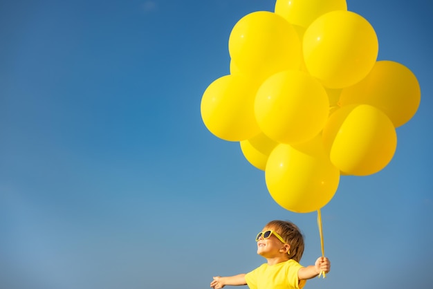 Criança feliz brincando com balões amarelos ao ar livre Criança se divertindo contra o fundo do céu azul de verão Liberdade e conceito de estilo de vida ativo