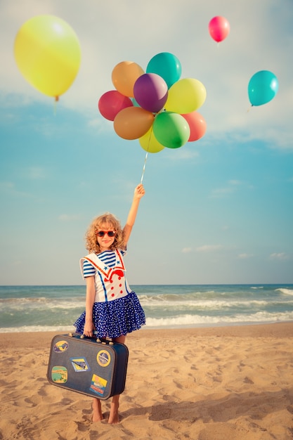 criança feliz brincando com balões coloridos e segurando uma mala na praia