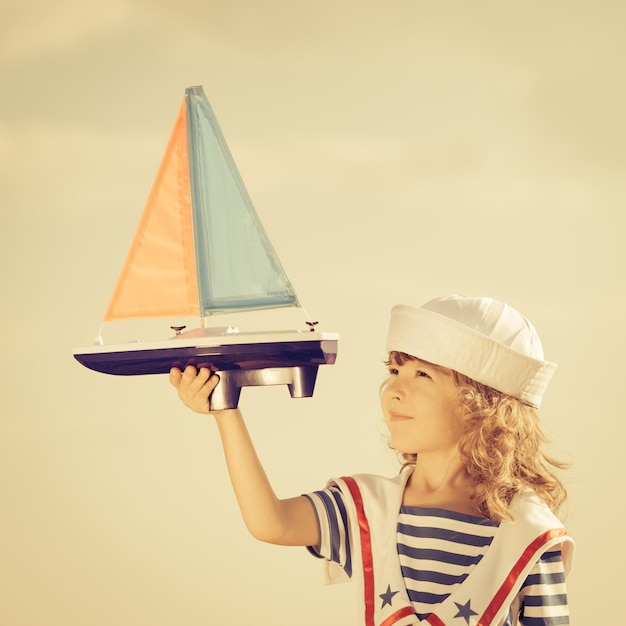 Criança feliz brincando com barco à vela de brinquedo contra o fundo do céu de verão Conceito de viagens e férias