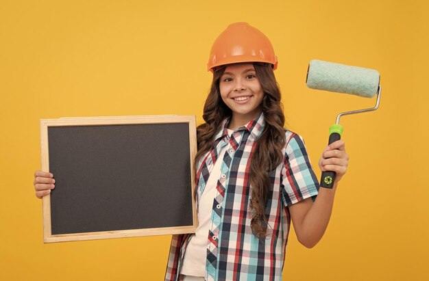 Criança feliz com cabelo encaracolado no capacete segura rolo de pintura e renovação do espaço de cópia do quadro-negro escolar