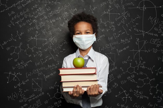 Criança feliz com máscara de proteção médica segurando livros e maçã verde no fundo do quadro preto