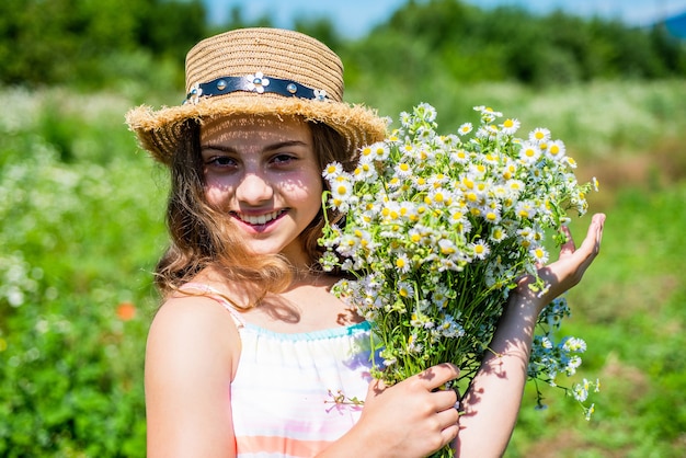 Criança feliz com sorriso de pele de bebê saudável com flores de camomila spa floral natural e cuidados com a beleza no chapéu de sol na paisagem ensolarada de verão, skincare.