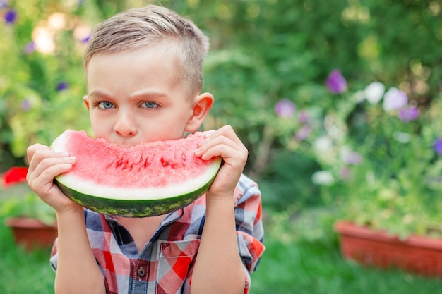 Criança feliz comendo melancia no jardim. As crianças comem frutas ao ar livre. Lanche saudável para crianças. Menino brincando no jardim, mordendo uma fatia de melancia.