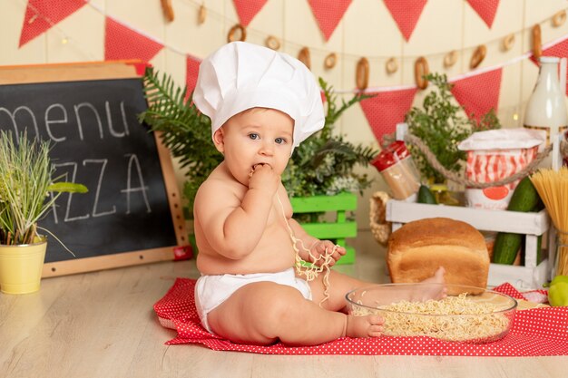 Criança feliz cozinhando de chapéu sentada na cozinha entre os produtos e comendo espaguete