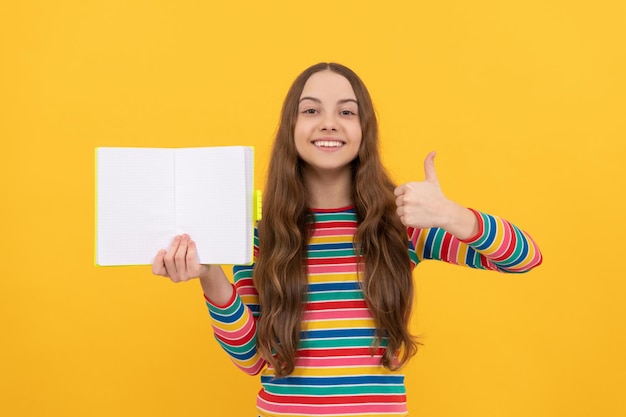 Criança feliz dá polegares para cima gesto com a mão segurando o livro aberto para cópia espaço fundo amarelo, satisfeito.