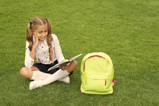 Criança feliz de uniforme com mochila lendo livro sentado na grama verde