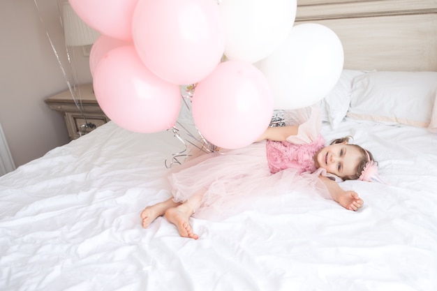 Criança feliz em um vestido festivo rosa com boné de aniversário deitada na cama branca aconchegante segurando balões banch