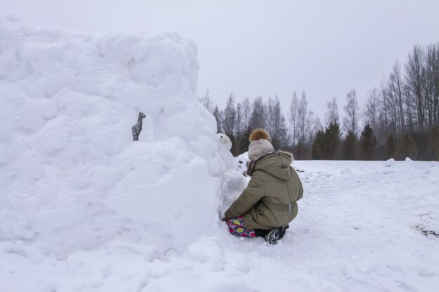 Criança feliz faz um boneco de neve em um campo coberto de neve na zona rural.