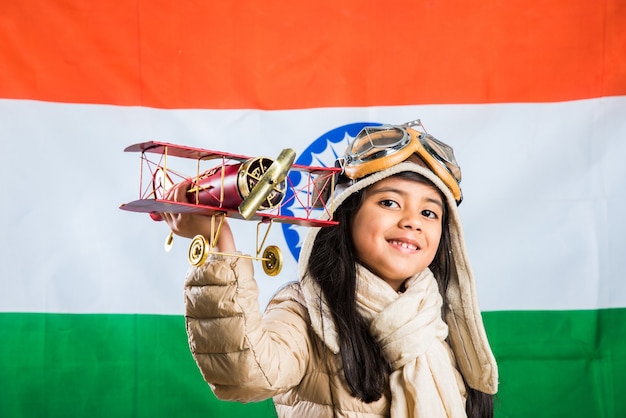 Criança feliz garota indiana ou asiática voando em um avião de brinquedo de metal ou saudando contra o fundo da bandeira indiana - criança e voando pelo país