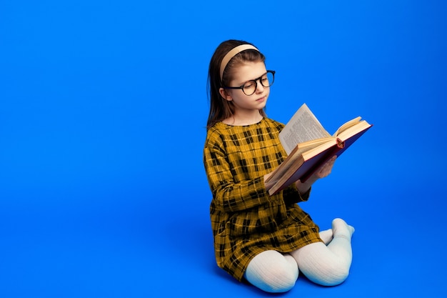 Criança feliz lendo um livro e sentada contra uma parede azul