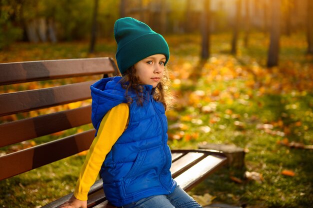 Criança feliz, linda menina com roupa quente colorida brilhante gosta do clima de outono, descansando em um banco de madeira do belo parque com folhas de bordo secas caídas ao pôr do sol. Infância, lazer outonal ao ar livre