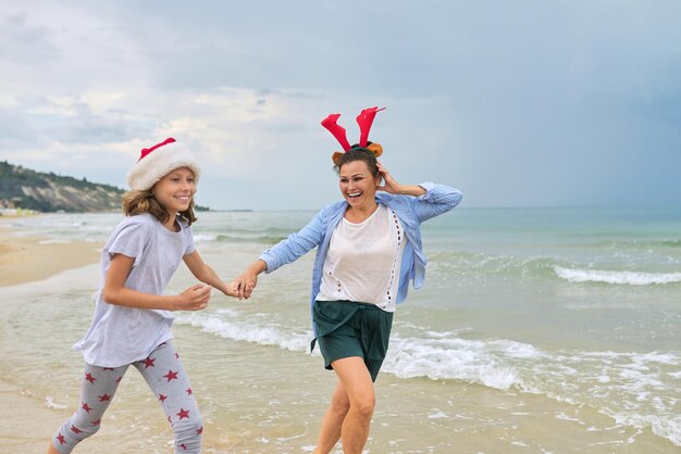 Criança feliz mãe e filha com chapéu de Papai Noel, se divertindo correndo ao longo da praia de mãos dadas. Família celebrando o Natal e o Ano Novo em um resort tropical, conceito de turismo de viagens de férias